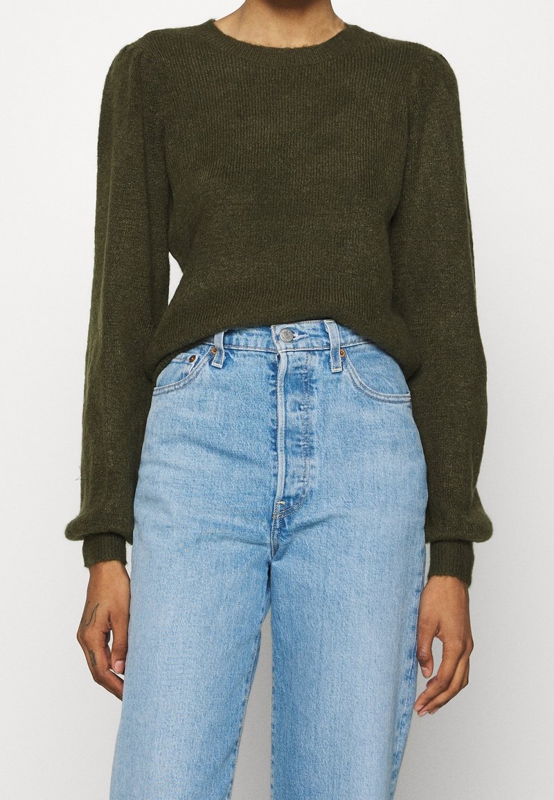 Personne portant un pull à manches longues vert olive rentré dans un jean taille haute bleu clair, debout devant un fond blanc.