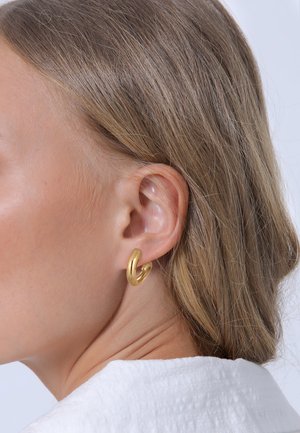 Goldenes Creolen-Ohrgerät mit einem gedrehten Design und einer glatten Textur. Das Ohrgerät ist am Ohr positioniert und teilweise von Haaren verdeckt.