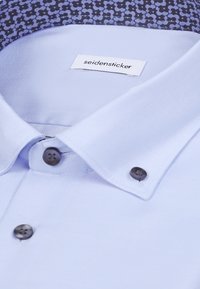 Chemise bleu clair avec boutons noirs et col intérieur à motif, étiquetée du nom de la marque "seidensticker."