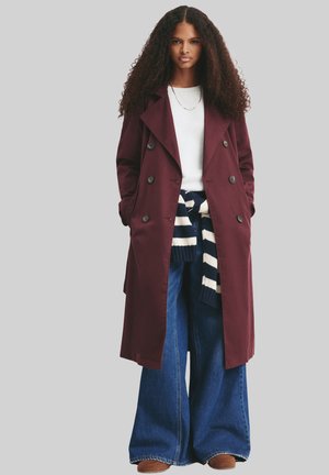 Femme aux longs cheveux bouclés portant un manteau bordeaux à double boutonnage, un pull blanc, un jean à jambes larges et un pull rayé noué autour de la taille.