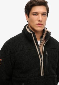 Czarna polarowa kurtka zapinana na zamek z wysokim kołnierzem, beżowymi akcentami i kieszenią na zamek na klatce piersiowej. Teksturowany materiał o miękkim wyglądzie.