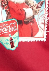 Rode stof met een vintage Coca-Cola ontwerp, inclusief een flessenlabel en een afbeelding van de kerstman, omgeven door decoratieve randen en tekst.