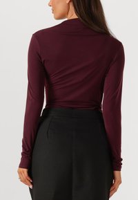 Langarmtop in bordeaux stof met een aansluitend ontwerp, voorzien van een gladde textuur en een subtiele drapering aan de achterkant. Gecombineerd met zwarte broek.