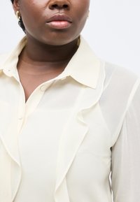 Κοντινό πλάνο ενός ατόμου που φοράει διάφανη κρεμ μπλούζα με λεπτομέρεια βολάν και χρυσούς κρίκους σκουλαρίκια.