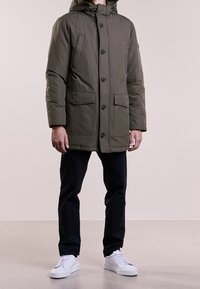 Oliwkowa, pikowana kurtka parka z kapturem, zapinana na guziki z przodu, z dwoma dużymi kieszeniami i regulowanymi mankietami, noszona na ciemnych spodniach i białych trampkach.