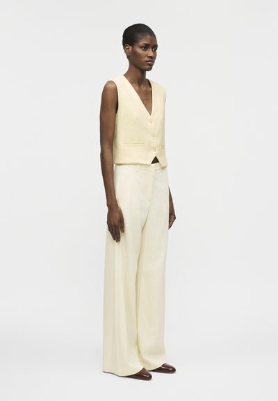 Γυναίκα που στέκεται, φορώντας αμάνικο μπεζ γιλέκο με κουμπιά και φαρδιά παντελόνια με ψηλή μέση σε μπεζ χρώμα, και καφέ παπούτσια.