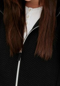 Lunghi capelli castani lisci su una camicia bianca con bottoni e un giubbotto nero con zip a trama.