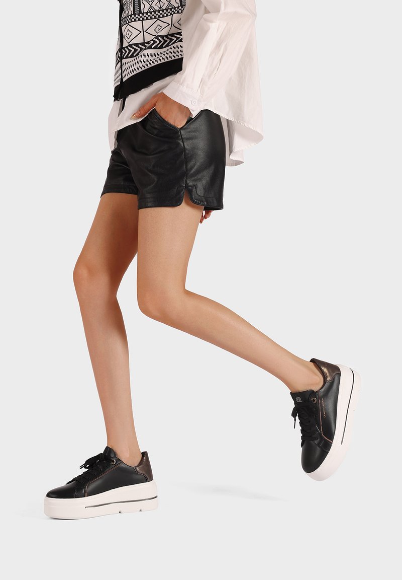 Osoba w czarnych skórzanych szortach, białej wzorzystej bluzce i czarnych platformowych trampkach, stojąca na jednej nodze na jednolitym tle.