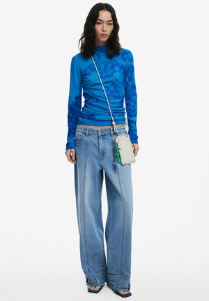 Blauw top met lange mouwen en bloemenpatroon, gecombineerd met loszittende lichtblauwe spijkerbroek en een kleine beige crossbodytas. Sandalen met hoge hak.