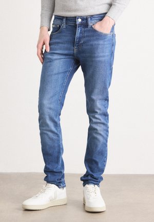 Persoon draagt slanke blauwe spijkerbroek, grijs shirt met lange mouwen en witte sneakers, staand op een lichte vloer tegen een effen achtergrond.