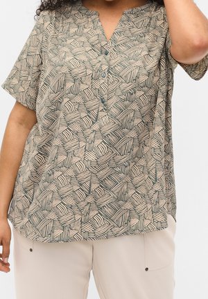 Loses Kurzarmhemd mit geometrischem beigem und grünem abstraktem Muster, durchgeknöpft, getragen mit hellbeigen Hosen.