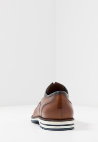 Chaussure en cuir marron à la forme arrondie, avec une texture lisse, des bords sombres contrastants et une semelle blanche avec des rayures bleues et rouges.