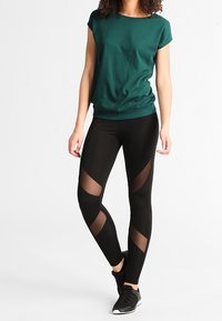 Leggings negros con secciones de malla recortadas y un diseño ajustado, combinados con una camiseta verde suelta. Cuello redondo simple, mangas cortas.