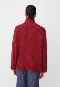 Czerwony sweter z wełny, z wysokim kołnierzem, luźnym krojem i ściągaczem u dołu. Tekstura wygląda na grubą, z nakrapianym wzorem w całej powierzchni.