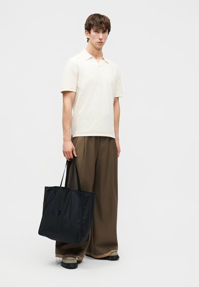 Jeune homme debout, portant un polo texturé ivoire à manches courtes, un pantalon large marron, des chaussures claires, et tenant un grand sac fourre-tout noir.