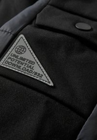 Zwarte textieljack met een glanzende afwerking, voorzien van een grijze driehoekige patch met de tekst "UNLIMITED POTENTIAL DOWNLOAD/932" en knoopdetails.