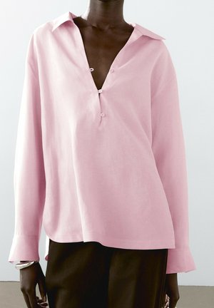 Persoon die een lichtroze blouse met lange mouwen draagt, met een diepe V-hals en knopen, gecombineerd met een donkere broek en een zilveren armband.