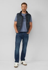 Opgevulde, donkergrijze vest met verticale stiksels, gedragen over een lichtblauw T-shirt met bedrukte tekst, in combinatie met blauwe jeans en witte sneakers.