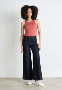 Mörkblå vida jeans i kombination med en figurnära korallfärgad linne med en liten logotyp, vita sneakers, mot en ljus bakgrund.