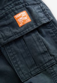 Sort stof med en tekstureret overflade, der har en orange etiket med teksten "SUPER DRY" og "CHAMPIONS 54 WORK CLOTHING" i hvid tekst.