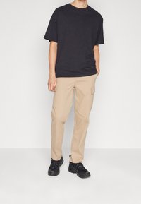 Persoon in een zwart t-shirt, beige cargobroek met zijzakken en zwarte sneakers, staande tegen een plain witte achtergrond.