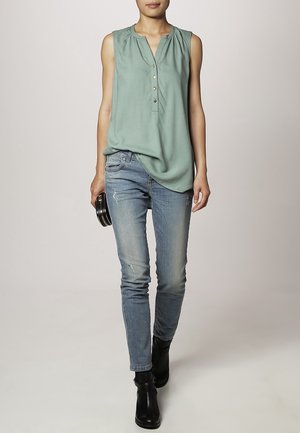 Femme portant un chemisier vert sans manches, un jean bleu déchiré, des bottines noires et tenant une petite pochette noire, marchant vers l'avant.