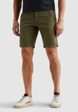 Mand iført olivengrønne knælange shorts, en sort skjorte og hvide sneakers, der står på en neutral baggrund.