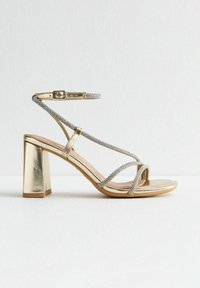 Golden high-heeled sandal featuring a chunky block heel, crisscross silver straps adorned with glitter, and an adjustable ankle buckle.