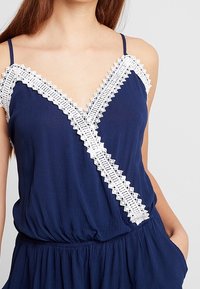 Navyblå ärmlös topp med v-ringning och vit spetsdetalj längs halsringningen och fram. Den har en r gathered midja och mjuk textur.