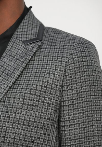 Blazer a cuadros grises con acentos negros, hombros estructurados, solapas de muesca y tejido texturizado. Primer plano del área del hombro y detalle del cuello.