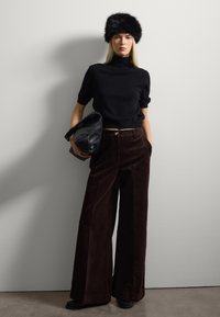 Top de punto negro de manga corta, pantalones de pana ancha marrón, gorro de piel negro, sosteniendo un bolso de piel negra texturizada, con un fondo gris claro.