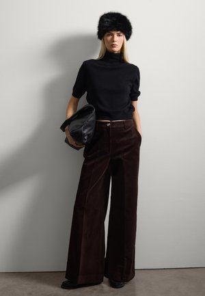 Top corto a maniche corte in maglia nera, pantaloni larghi in corduroy marrone, cappello di pelliccia nera, con in mano una borsa in pelle nera strutturata, su uno sfondo grigio chiaro.
