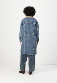 Veste en denim à coupe longue, forme structurée et couture visible. Dotée d'un col doux contrasté et de poches arrière. Associe avec un pantalon en denim.