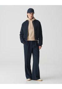 Marineblauer Bomberjacke, beiger Kapuzenpullover, marineblaue weit geschnittene Hose und marineblauen Hut. Baumwollmischung, lockere Passform, mit glatter Textur und klassischem Design.
