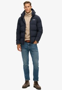 Marineblåt puffjakke med hætte, med et logo på brystet, parret med en beige turtleneck, blå jeans og brune snørebod.