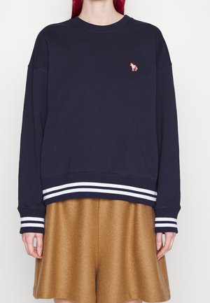 Personne portant un sweat-shirt bleu marine avec des poignets et un ourlet rayés blancs, avec un petit emblème de cheval multicolore, associé à un short couleur camel.