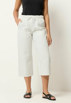 Kobieta ubrana w białe spodnie trzy czwarte z cienkimi czarnymi pionowymi paskami, czarną bluzkę bez rękawów i czarne płaskie sandały, stojąca z rękami w kieszeniach.