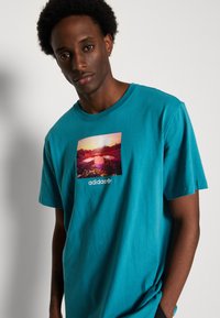 Teal puuvillane T-särk, millel on ristkülikukujuline graafiline trükis päikeseloojangust maastikust ja Adidas logo valge värviga allosas keskpunktis.