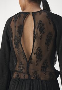 Kobieta w czarnej koronkowej sukience z przezroczystym panelem z kwiatowej koronki na plecach i małym okrągłym zapięciem przy dekolcie.