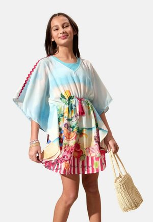 Fată îmbrăcată într-o rochie de plajă colorată cu modele de fructe și dungi, ținând o geantă de paie împletită, zâmbind pe un fundal simplu.