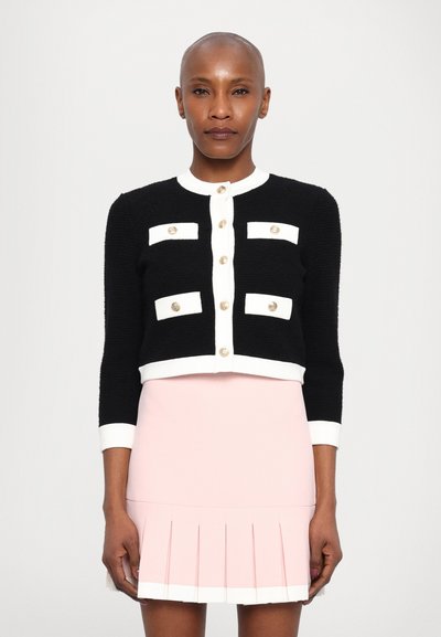Cardigan noir court avec des accents blancs, des boutons dorés et un tissu texturé ; associé à une jupe plissée rose pâle.