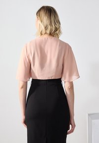 Kobieta z krótkimi blond włosami, ubrana w przezroczystą różową bluzkę z rękawami do łokcia i czarną spódnicę z wysokim stanem, widziana od tyłu.