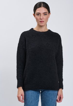 Woman with dark hair in a black textured sweater and blue jeans standing against a plain light background, facing forward.