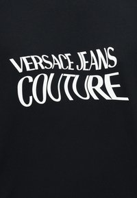 Tela negra con texto blanco distorsionado que dice "VERSACE JEANS COUTURE" en letras mayúsculas.