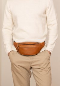 Man draagt beige broek en witte trui met een bruine leren heuptas om de taille vastgemaakt, handen in de zakken.