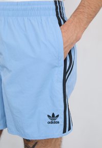 Világos kék Adidas rövidnadrág fekete oldalsó csíkokkal és logóval. Könnyű anyagból készült; rugalmas derékkal és oldalsó zsebekkel rendelkezik.