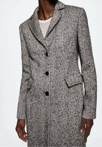 Grijze visgraatmantel met een ingesneden kraag, voorzien van zwarte knopen en twee voorzakken. Getextureerde wollen stof voegt diepte toe.