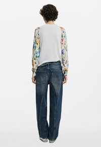 Haut blanc avec des manches à motifs floraux colorés, associé à un jean bleu foncé à coupe décontractée avec poches arrière et ourlet brut.