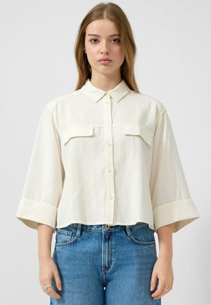 Frau mit glattem, hellbraunem Haar, die ein lockeres, cremefarbenes Hemd mit Knöpfen trägt, dessen dreiviertel-lange Ärmel hochgekrempelt sind, und blaue, hochtaillierte Jeans.