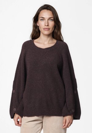 Femme portant un pull en maille ample marron foncé à manches larges avec détails de boutons, associé à un pantalon beige clair sur fond blanc.